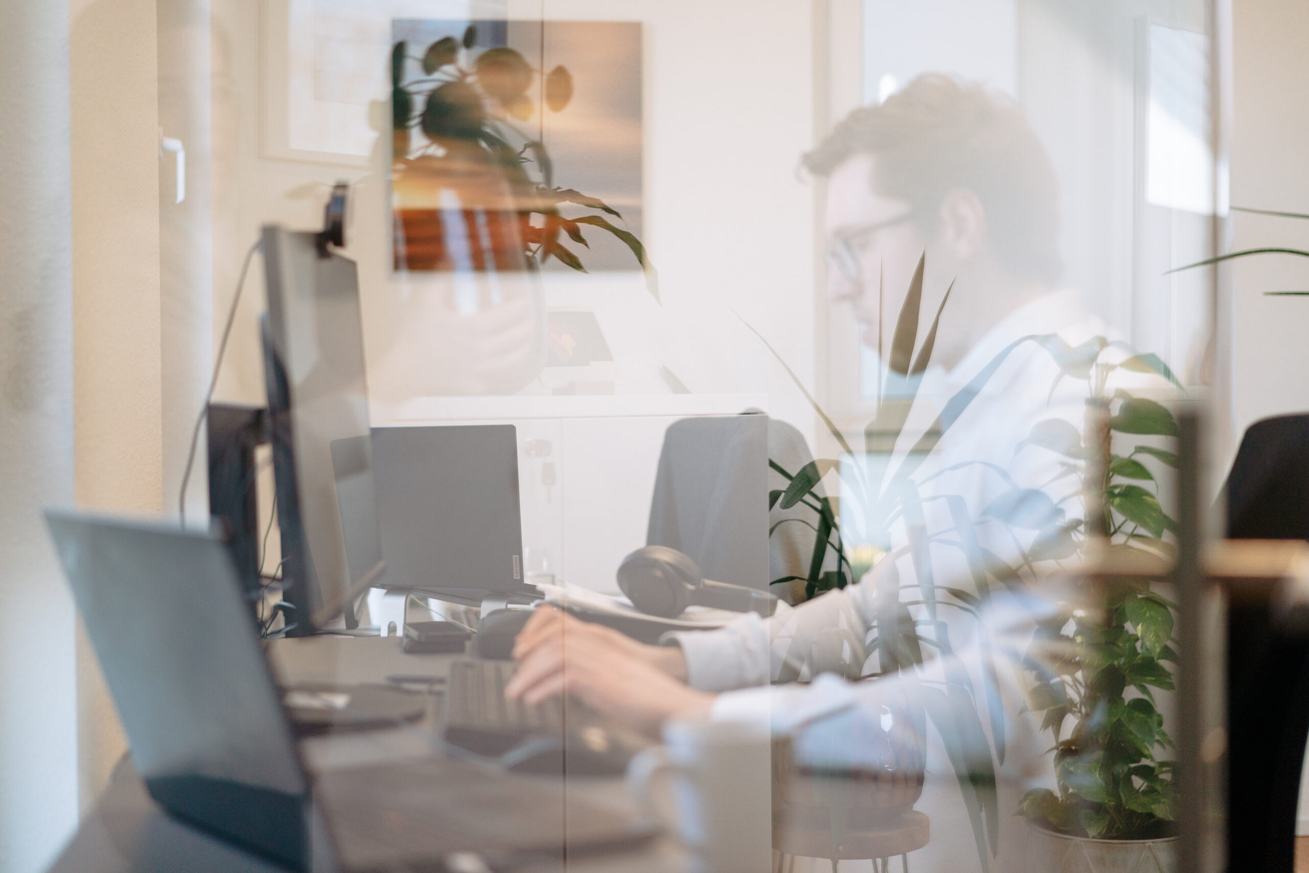 Ein Mitarbeiter mit Brille und weißem Hemd sitzt an einem Schreibtisch und arbeitet konzentriert am Computer. Die Aufnahme entstand durch eine Glasscheibe, in der sich Pflanzen, Fenster und Teile der Büroeinrichtung spiegeln. Mehrere Bildschirme, ein Laptop und weitere Arbeitsutensilien prägen die moderne Büroumgebung. Das natürliche Licht sorgt für eine helle und angenehme Arbeitsatmosphäre.