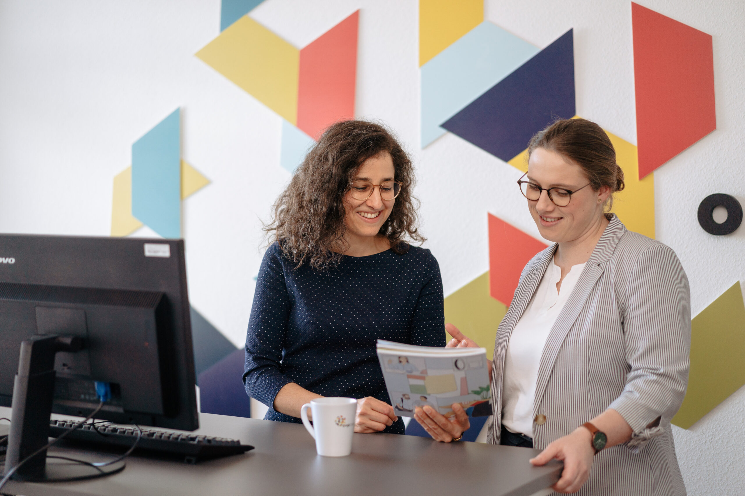 Zwei Beraterinnen stehen an einem Schreibtisch und besprechen gemeinsam Unterlagen. In moderner Büroumgebung tauschen sie sich konzentriert aus – sachlich, professionell und kooperativ.