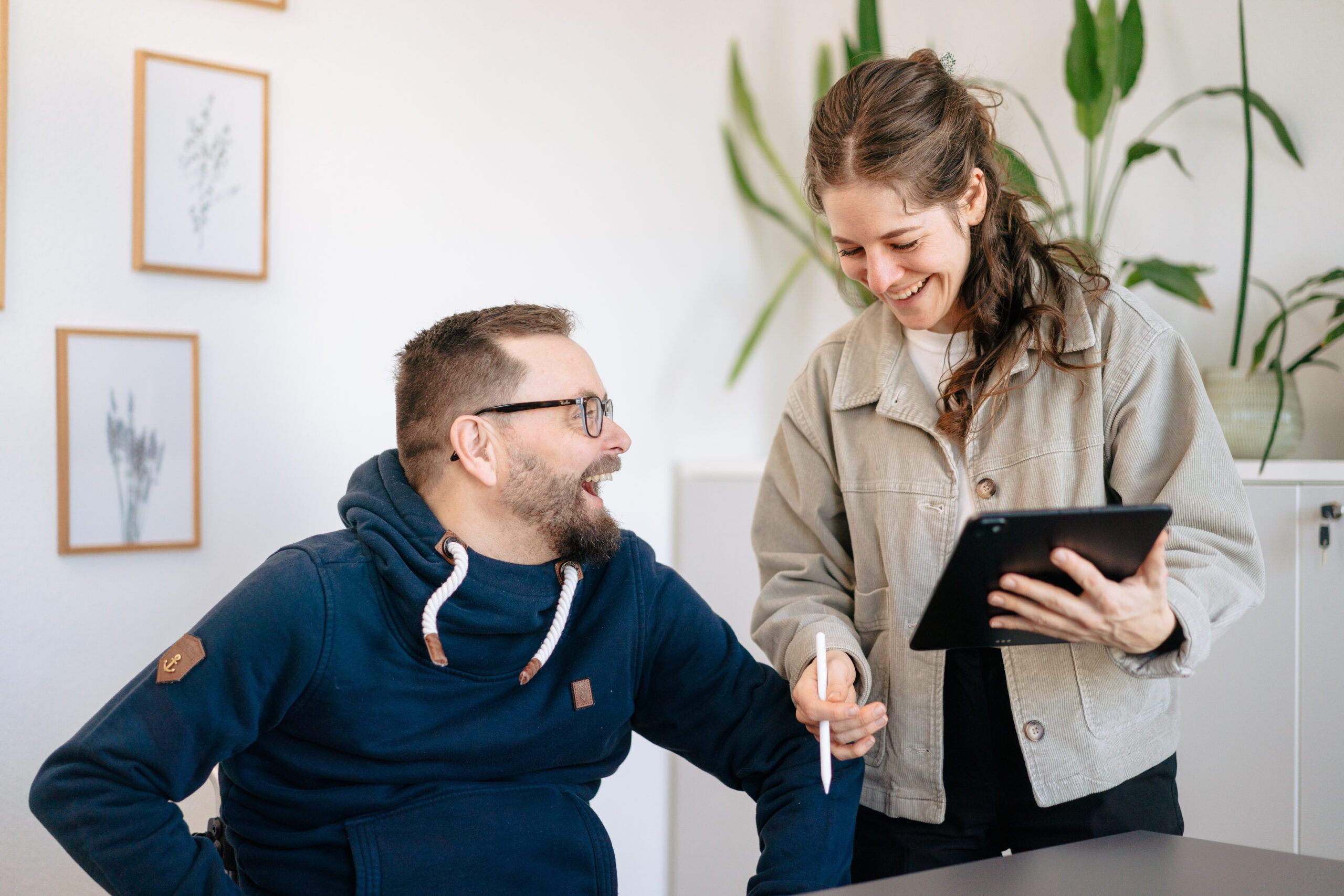 Praktikum Lernen im Team, eigene Ideen einbringen, Verantwortung übernehmen, ein Praktikum bei uns bietet echte Einblicke und Entwicklung, zwei Personen lachen in heller Arbeitsumgebung, eine Person steht mit Tablet in der Hand