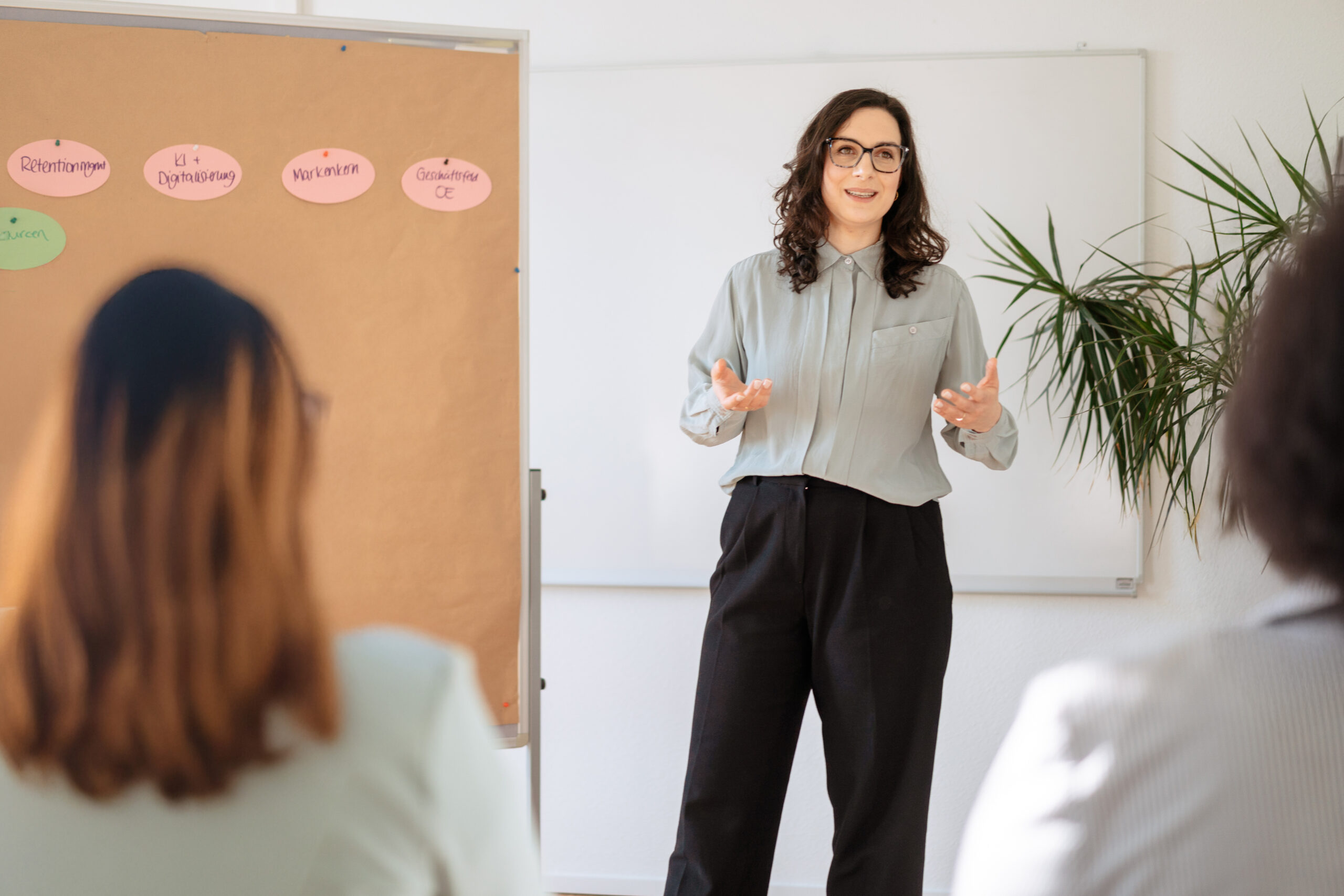 Eine Frau spricht vor einer kleinen Gruppe, hinter ihr Themenkarten an einer Pinnwand, vermitteln von Softskills an eine Gruppe von Trainingsteilnehmer:innen