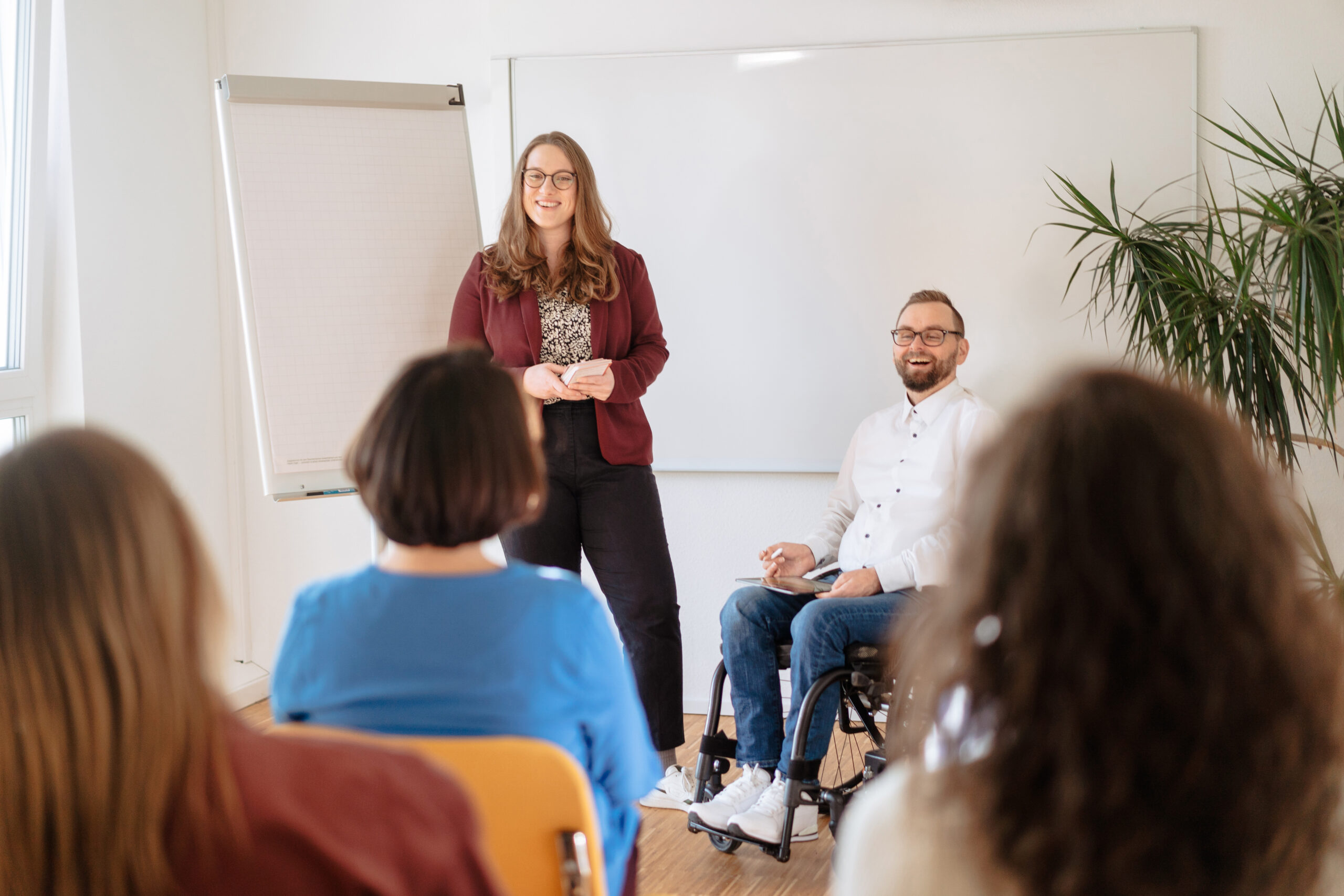Zwei Personen leiten ein Training, stehen und sitzen vor einer Gruppe in hellem Raum, vermitteln von Softskills an eine Gruppe von Trainingsteilnehmer:innen