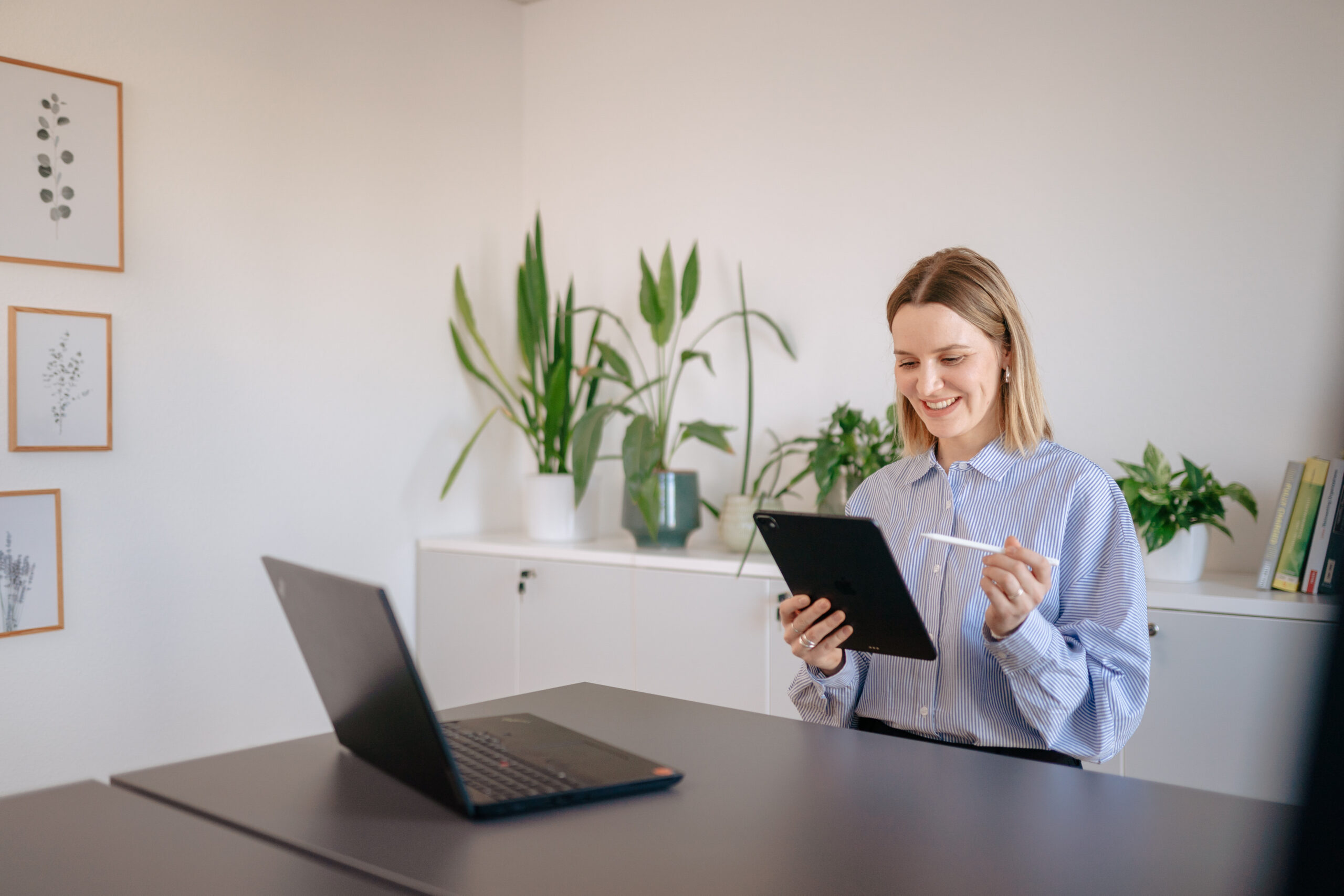 Überblick MiWay Motivations- und Werteanalyse Eine Frau sitzt am Schreibtisch, arbeitet lächelnd mit Tablet und Stift in heller Umgebung, Reflexion von motivierenden und die motivierenden beruflichen Werten