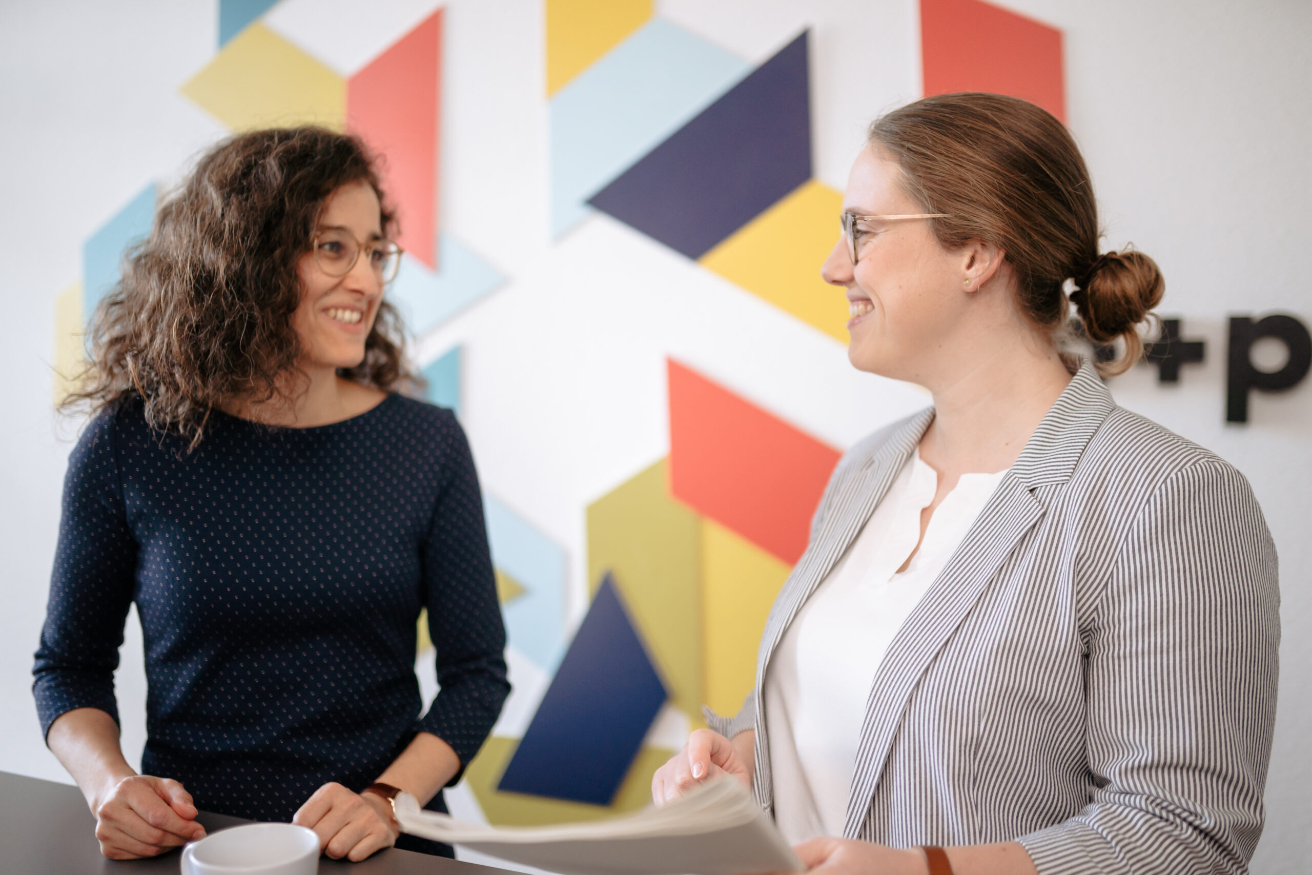 Überblick MyCulture Kulturanalyse Zwei Frauen im Gespräch, lächeln sich an, bunte geometrische Wand im Hintergrund, freundliche Atmosphäre, Austausch über Anforderungen an Unternehmenskultur