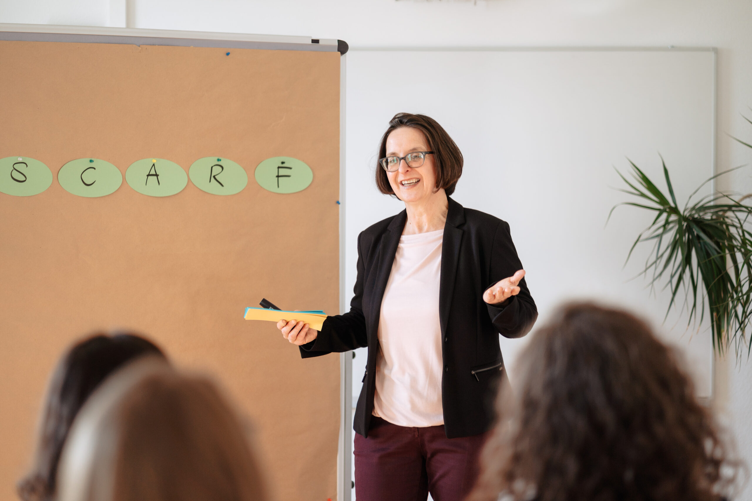 Eine Frau steht lächelnd vor einer Pinnwand, präsentiert das SCARF-Modell vor einer Gruppe, Vermitteln von Inhalten Teilnehmende aus unterschiedlichen Organisationen