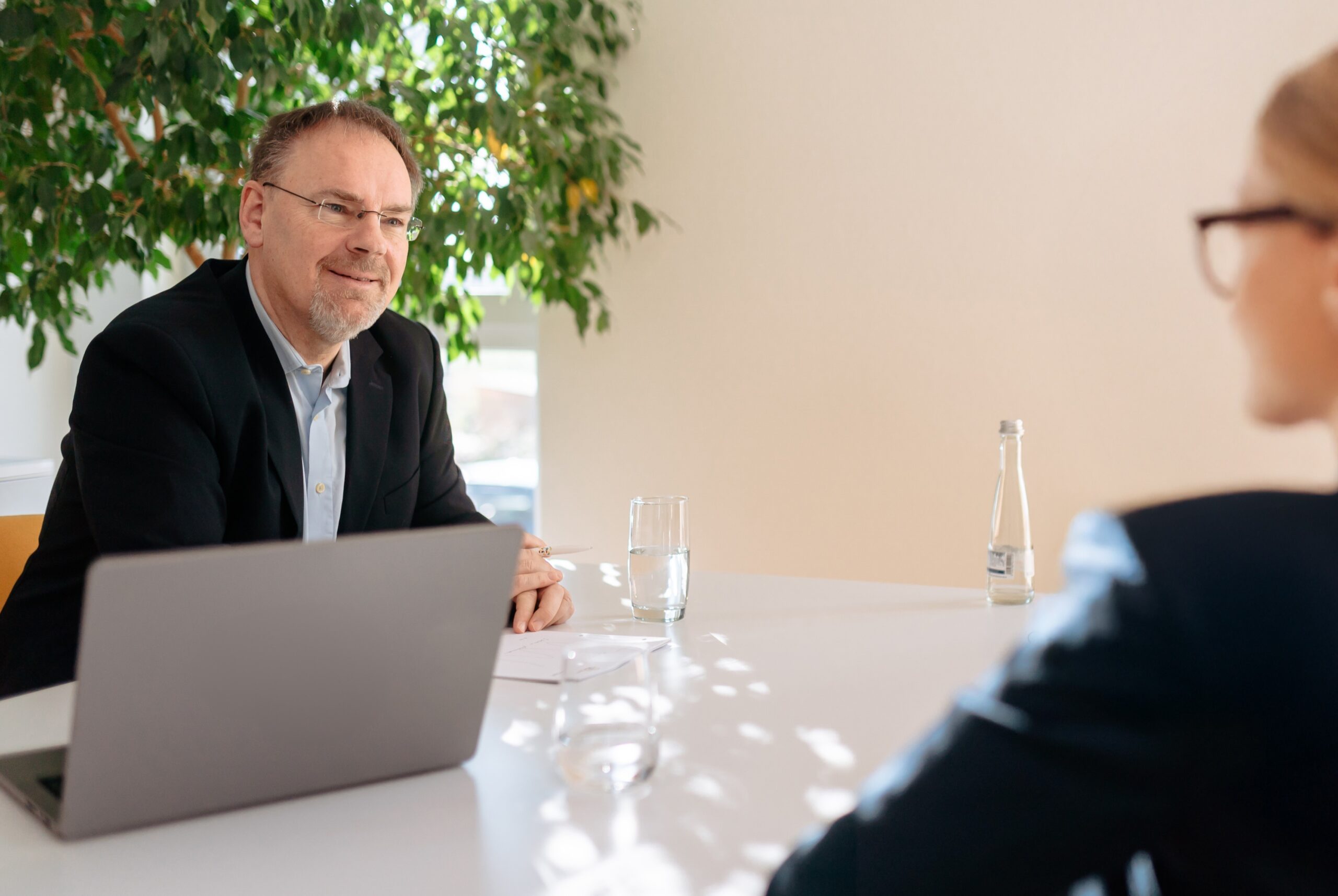 Ein Mann sitzt am Schreibtisch einer Frau gegenüber, helle, freundliche Büroatmosphäre, ein Laptop steht auf dem Tisch, Begleitung einer umfassenden Eignungsdiagnostik mit verschiedenen Elementen