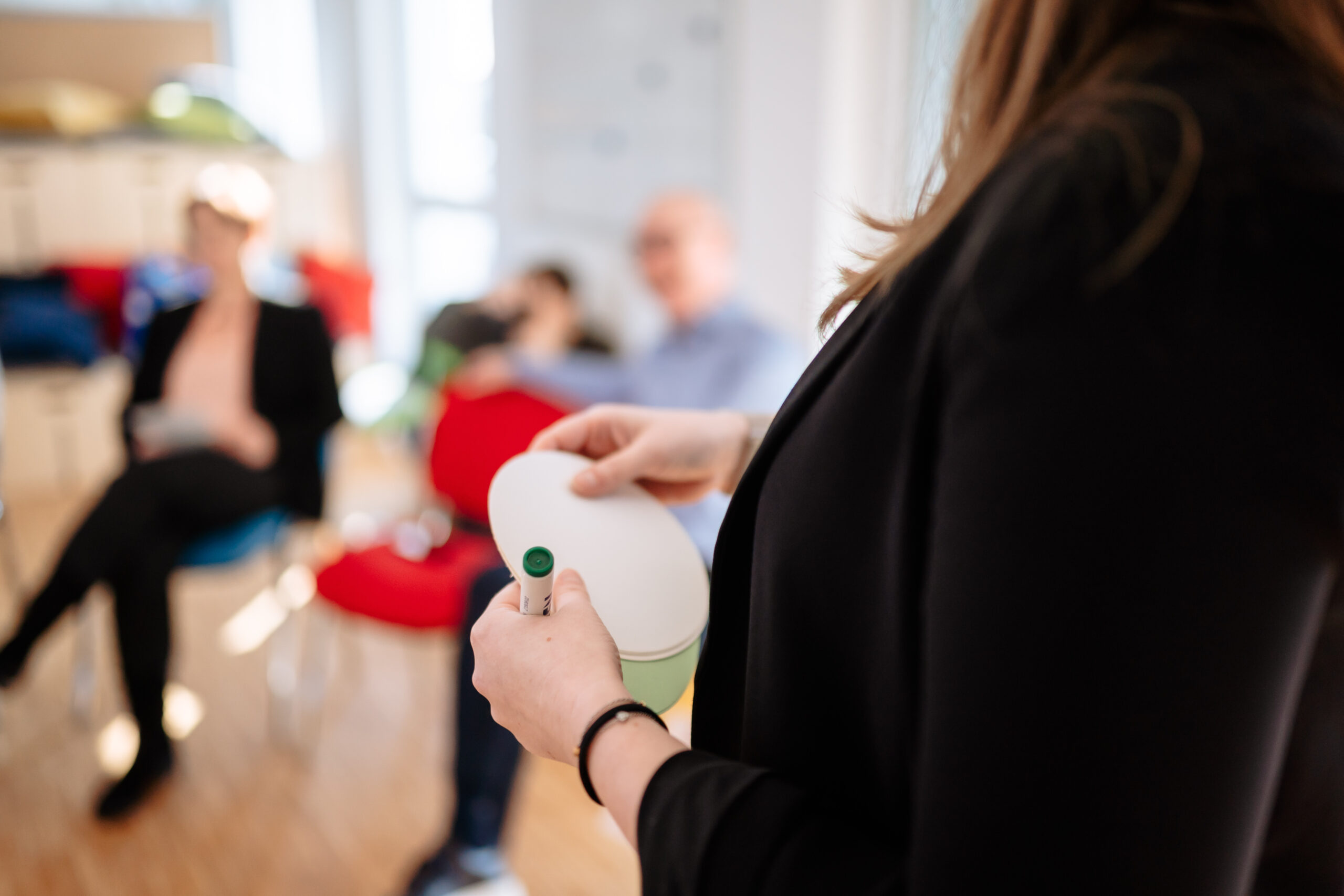 Unser Ansatz Kompetenzmanagement Eine Frau steht vor einer Gruppe und hält ein Moderationskärtchen, aufmerksames zuhören, Verstehen der unterschiedlichen Rollen, die in der Organisation existieren, Festlegen von unterschiedlichen Anforderungsniveaus