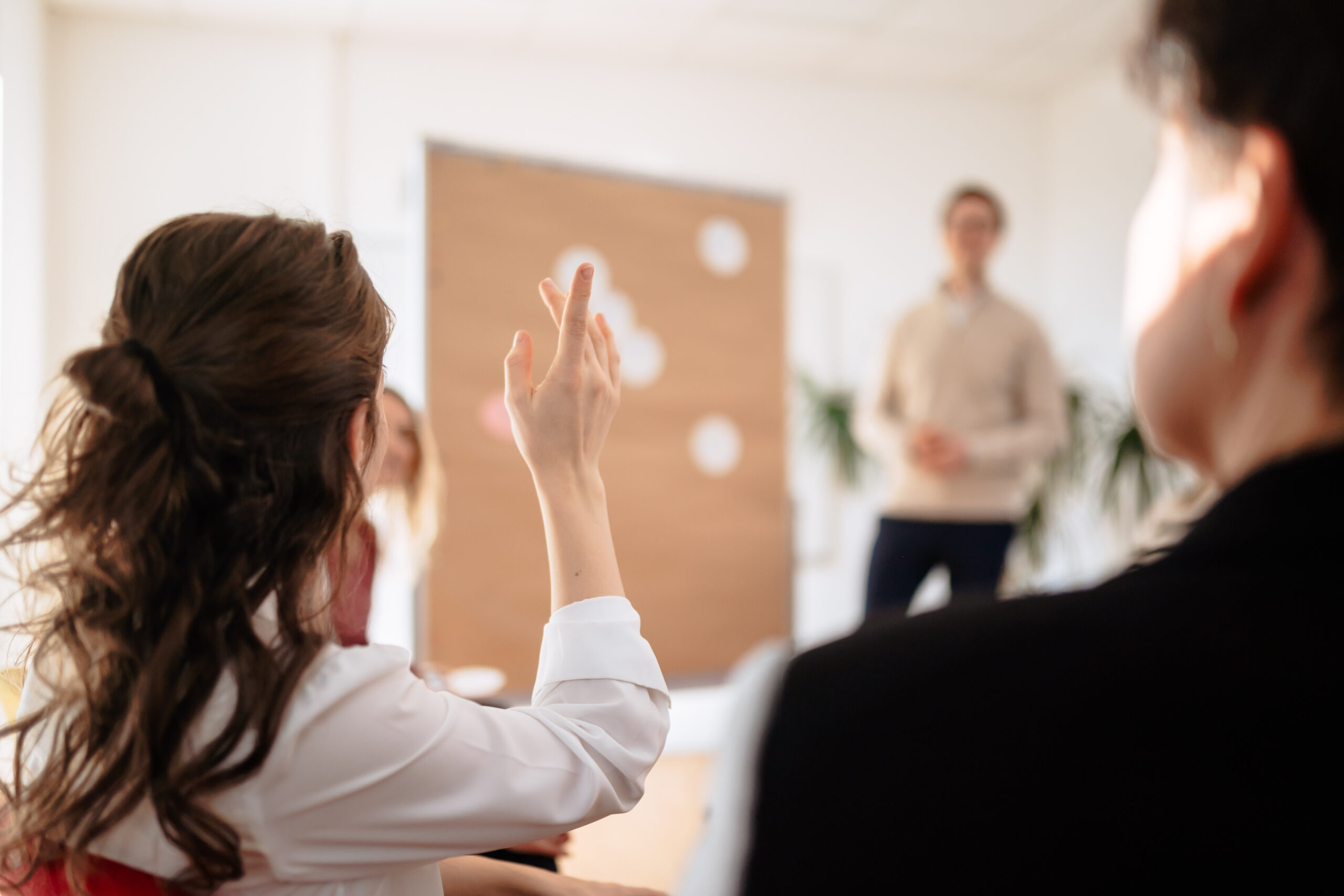 Eine Frau im Vordergrund meldet sich, ein Mann im Hintergrund steht an einer Metaplanwand, klaren Fokus auf Reflexion eigener Stärken und Entwicklungsfelder, Feedback und innere Entwicklung