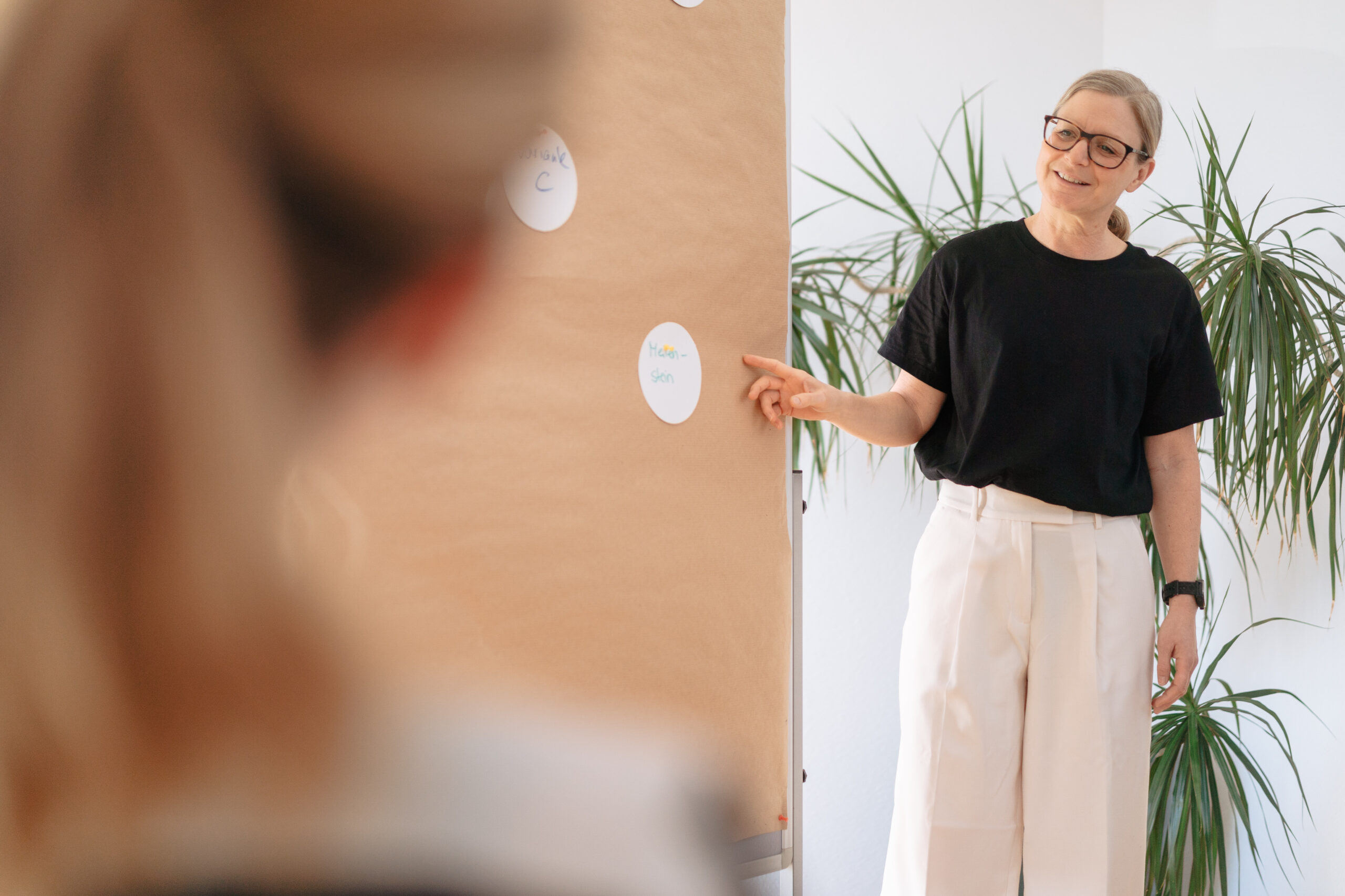 Unser Ansatz Retention Management Eine Frau steht vor einer Metaplanwand, im Hintergrund eine Pflanze, helle Atmosphäre, Betrachtung des Einstellungsprozesses, der Einarbeitung neuer Mitarbeitender und Möglichkeiten zur langfristigen Weiterentwicklung