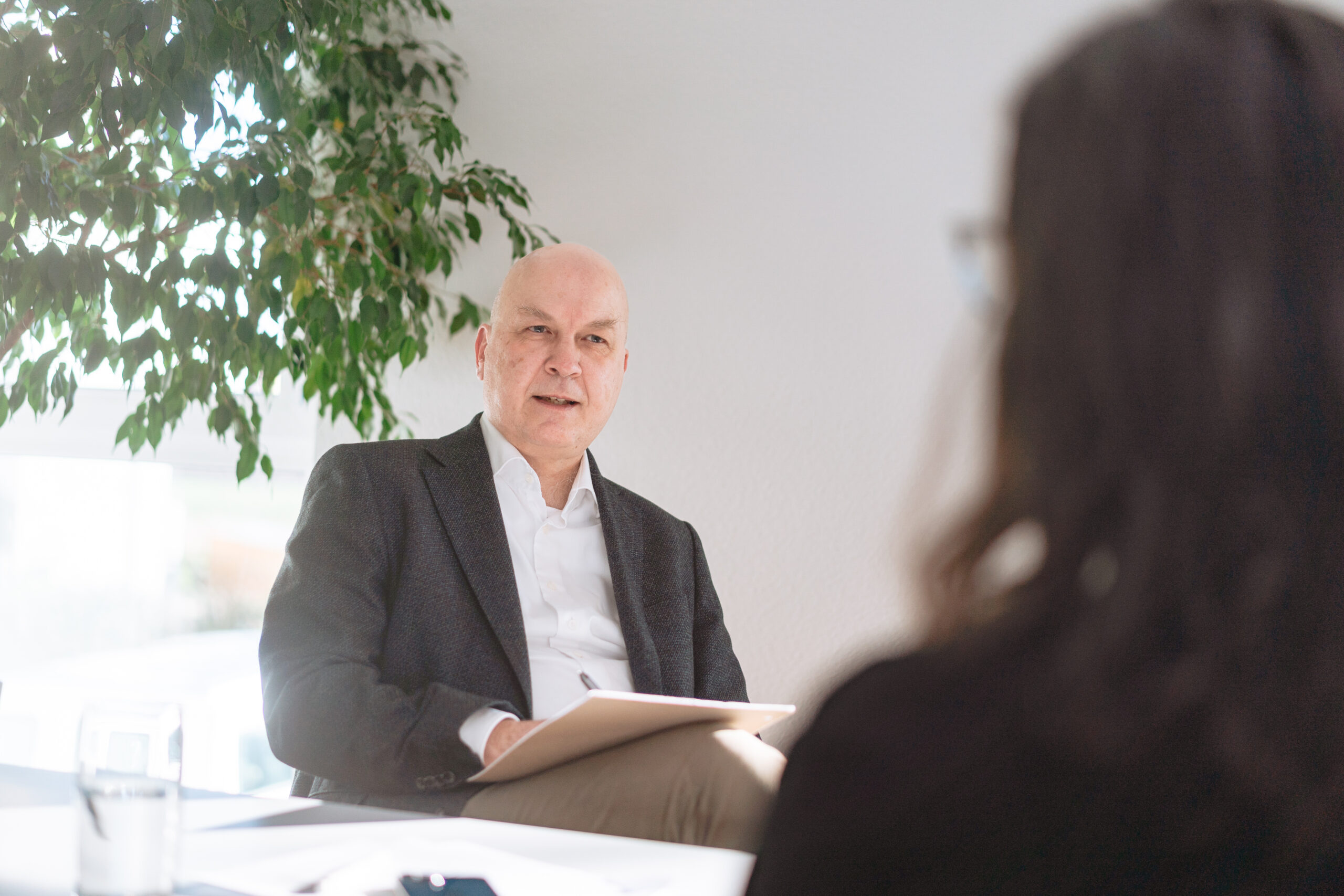 Unser Ansatz Talent Management Ein älterer Mann hört aufmerksam zu, macht sich Notizen, im Vordergrund eine Frau, helle Atmosphäre mit einem Pflanze, Nachfolgeplanung, Karriereplanung, frühzeitig Talente entdecken
